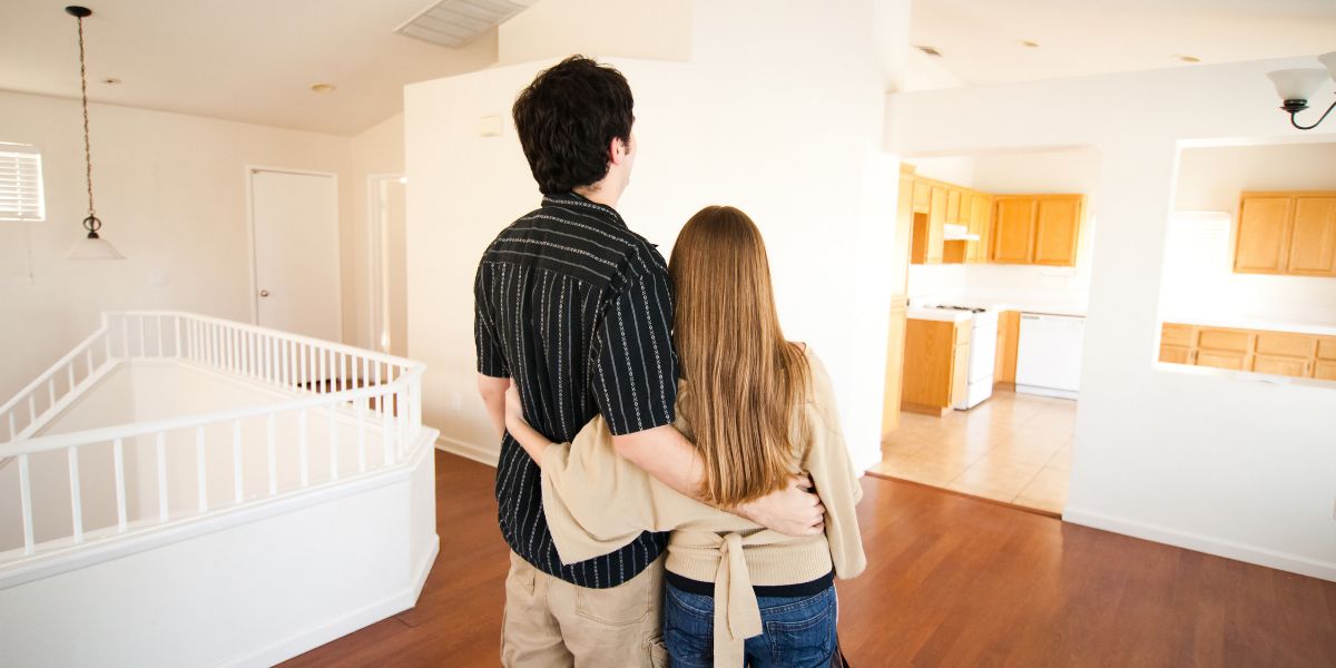 Couple standing arm in arm, looking at an empty room in a house with wood floors and white walls.