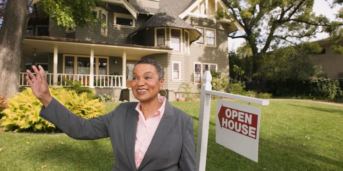 Smiling woman waving in front of a house with an 'Open House' sign in the yard.